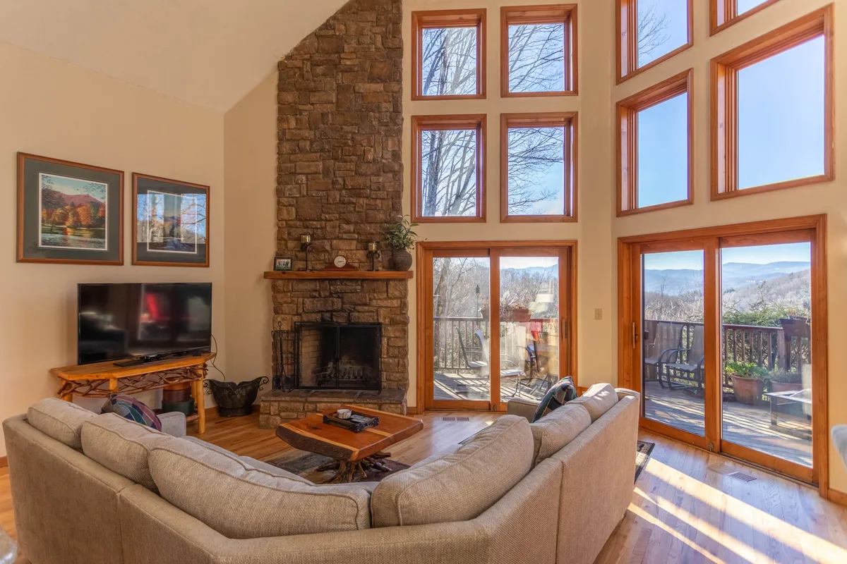 A spacious great room with a prominent stone fireplace serves as the focal point, surrounded by an expansive sectional sofa and a large wooden coffee table. Floor-to-ceiling windows flood the space with natural light and showcase scenic mountain views, enhancing the room's airy, open feel. The overall mood is warm and welcoming, ideal for gatherings and relaxation.