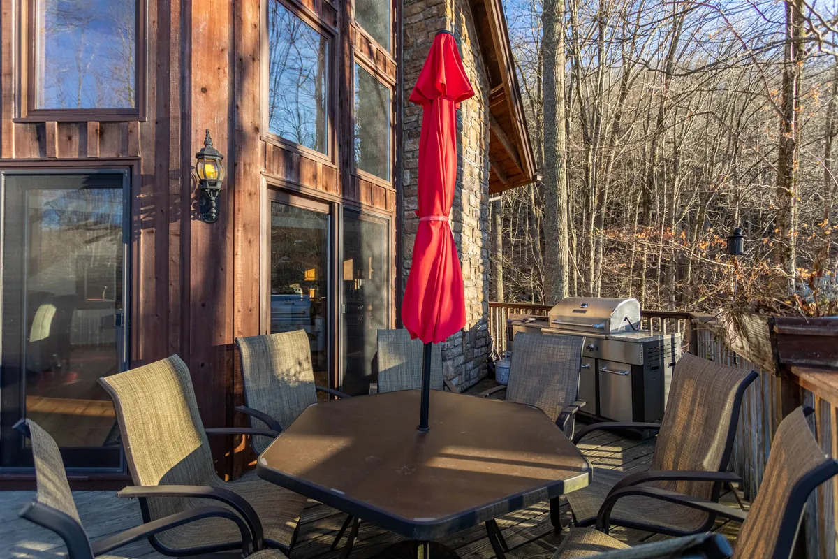 An outdoor living area featuring a red umbrella, generous outdoor seating, a grill, stone wall, and large windows that hint at the indoor spaces beyond. The area is designed for relaxation, outdoor dining, and social gatherings, all set within natural surroundings.