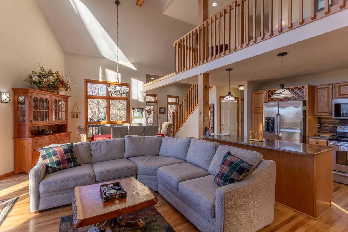 A view of the open-concept living, dining, and kitchen areas, featuring a large sectional sofa, a wooden dining table perfect for group meals, and an open staircase. The kitchen’s countertop is visible, reinforcing the sense of a unified, communal space designed for socializing, cooking, and relaxing together.