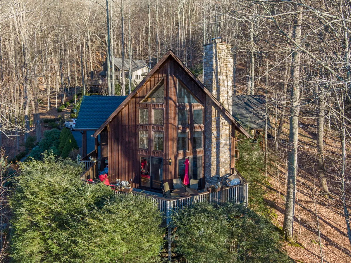 This image features a cozy wooden cabin with a prominent stone chimney, large windows, and a deck furnished with chairs. The setting is surrounded by trees, creating a tranquil and inviting atmosphere that blurs the line between indoor comfort and outdoor beauty.