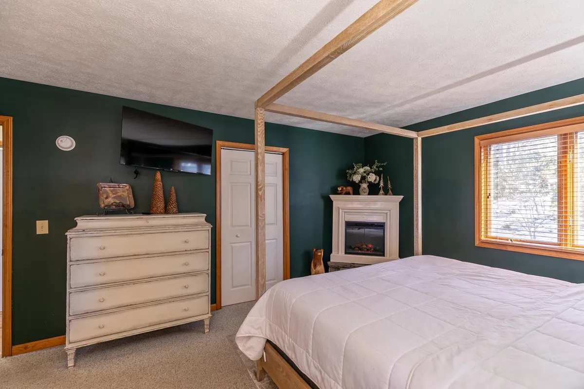 A serene bedroom with a four-poster bed and a fireplace, positioned next to a window with a view. The space exudes comfort and warmth, ideal for relaxing after a day of winter activities or summer exploration. The window view subtly suggests the changing scenery outside.