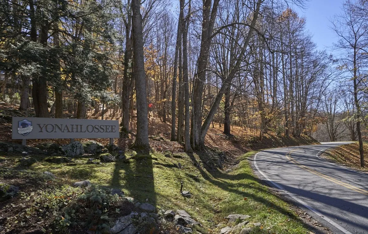The image features the prominent YONAHLOSSEE sign at the entrance to the resort, set along a winding road flanked by trees with vibrant autumn foliage. The scene feels welcoming and hints at the resort's natural beauty and sense of arrival.