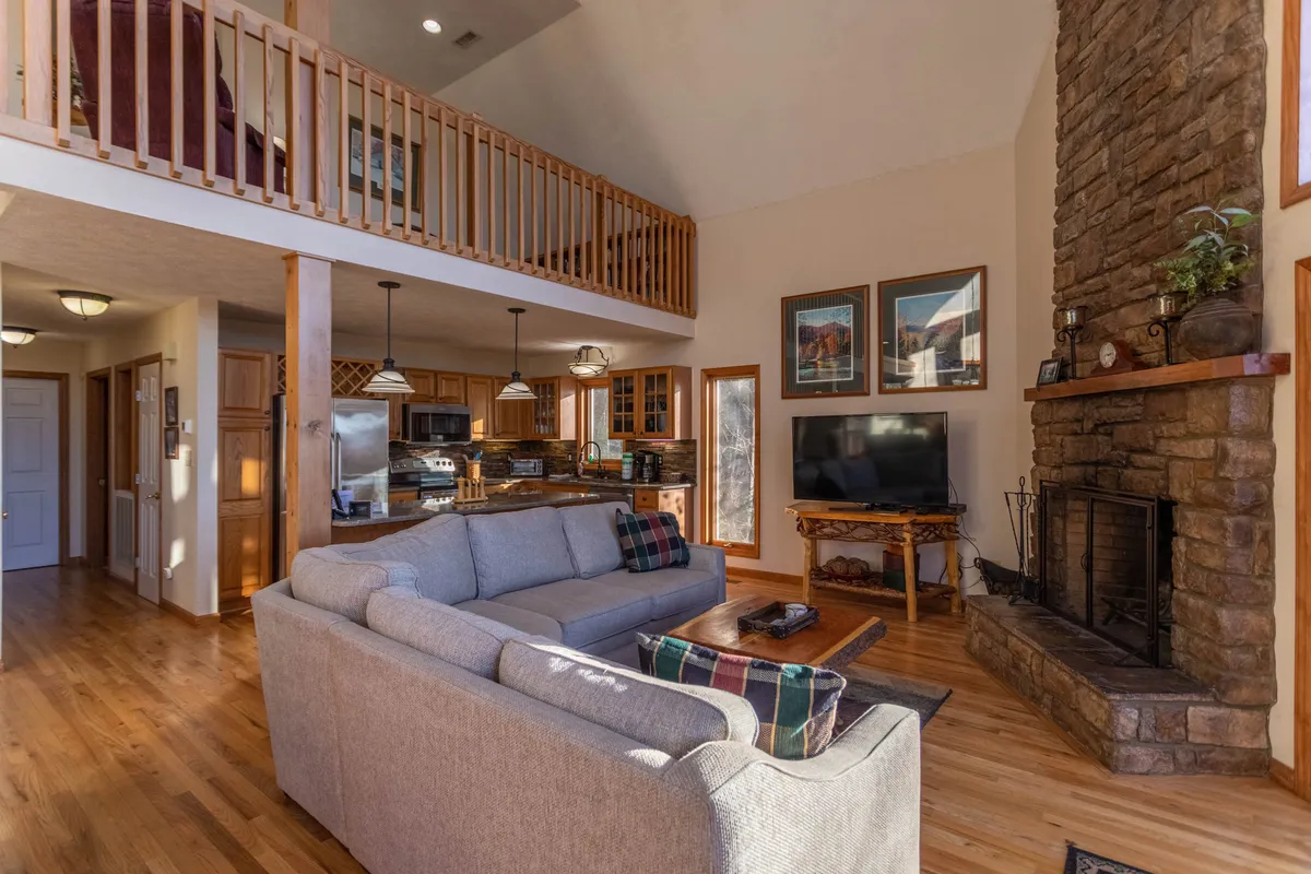 A welcoming living room featuring a stone fireplace, comfortable sofa, and expansive windows that allow natural light to flood the space. The open loft area and views through the large windows hint at the natural beauty outside, inviting guests to relax and observe the seasons.