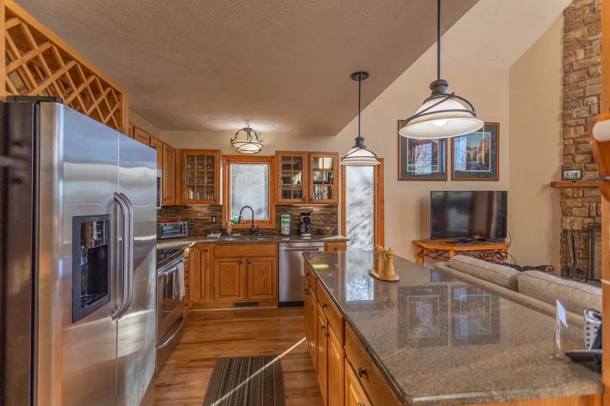 A spacious, homey kitchen anchored by a central island and surrounded by stainless steel appliances, warm wooden cabinetry, and a cozy stone fireplace. Subtle wall art completes a welcoming atmosphere tailored for group cooking, entertaining, and shared meals.