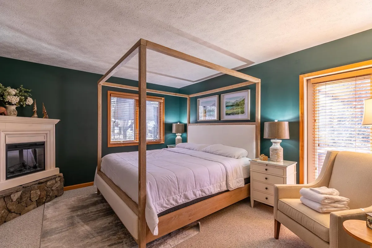 A spacious and inviting bedroom featuring a stately four-poster bed, elegant fireplace, comfortable armchair, and tasteful framed artwork. The natural tones and soft lighting evoke a sense of tranquility and comfort, perfect for restful nights or cozy mornings in bed.
