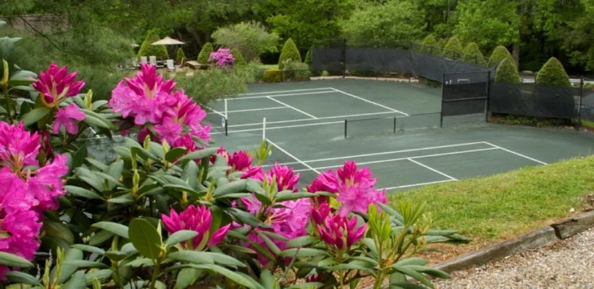 An outdoor tennis court framed by vibrant pink flowers and greenery. The scene hints at matches being played, while the surrounding landscape underscores a lively yet refreshing resort atmosphere.