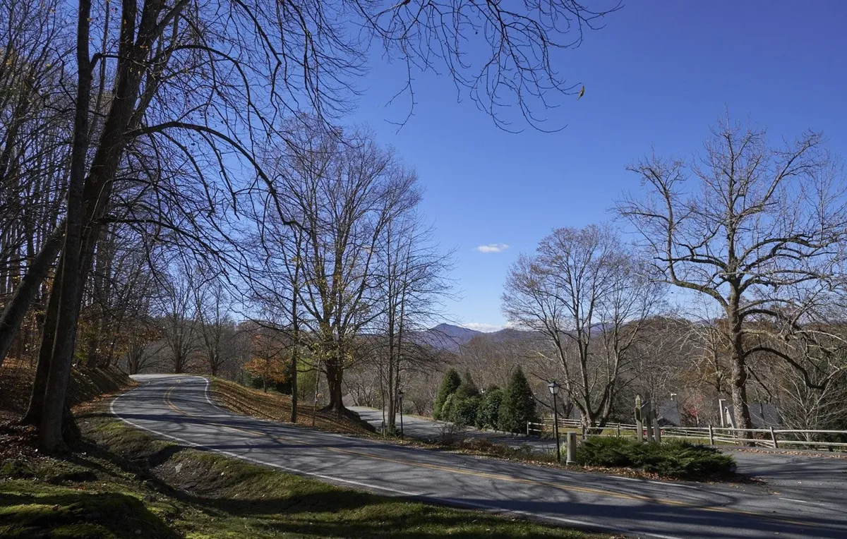 This exterior scene captures a winding road flanked by bare trees, leading toward a distant mountain backdrop. The landscape is serene and open, evoking the sense of adventure and scenic exploration that defines North Carolina's High Country. The composition highlights the natural setting and the allure of hitting the road to discover nearby trails, waterfalls, and the Blue Ridge Parkway.